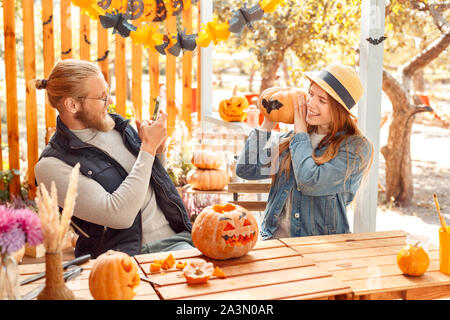 Halloween Preparaton concetto. Coppia giovane seduto a tavola all'esterno rendendo jack-o'-lanterna uomo prendendo foto di donna in posa con la zucca gioiosa Foto Stock