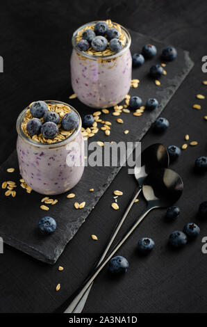 Colazione sana nozione. Vasetti di yogurt fatto in casa con mirtilli e avena su sfondo nero. Verticale. Foto Stock