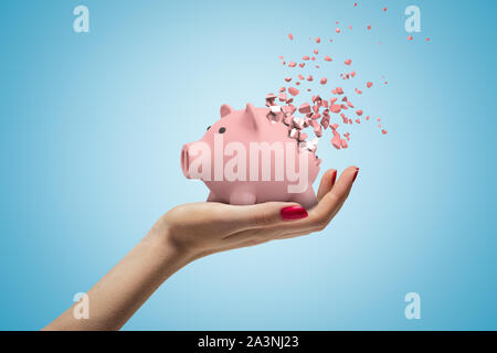 Close-up di donna di mano rivolto verso l'alto e mantenendo carino rosa salvadanaio che ha iniziato a disintegrarsi in pezzi su luce-sfondo blu. Foto Stock