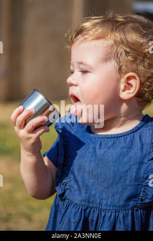 Piccola bambina di 1 anni in un vestito blu, aprire bocca linguetta sporgente, vuole il bambino di bere da un bicchiere, closeup ritratto. Ragazza caucasica con Foto Stock