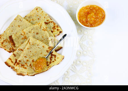 Crepes piegato in quattro sulla piastra bianca e marmellata di arance amare con cucchiaio su sfondo bianco. Vista superiore Foto Stock