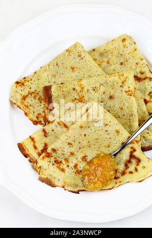 Crepes piegato in quattro sulla piastra bianca e marmellata di arance amare con cucchiaio su sfondo bianco. Vista superiore Foto Stock
