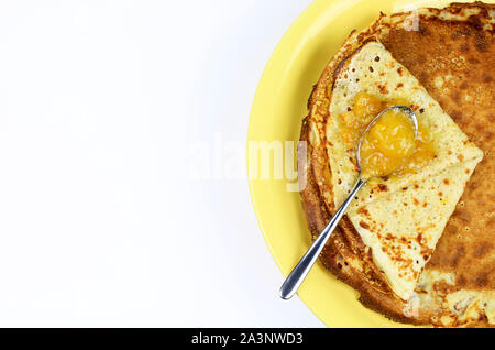 Crepes piegato in quattro sulla piastra di colore giallo e marmellata di arance amare con cucchiaio su sfondo bianco. Vista superiore Foto Stock