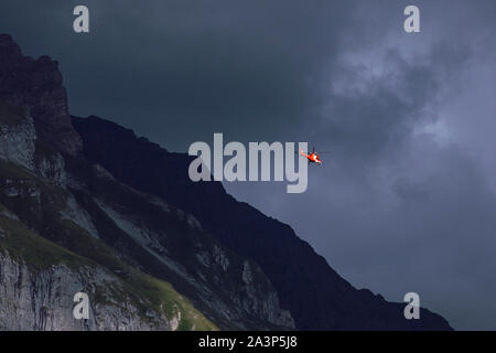 Elicottero rosso sul cielo nuvoloso sfondo. Grande oscura montagna. Salvataggio in elicottero nelle montagne rocciose, crepuscolo. Swiss montagne alpine. Svizzera Foto Stock