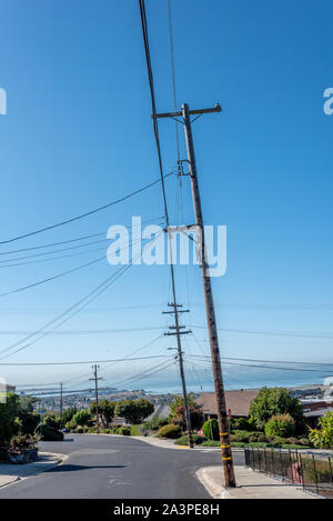 Alti pali del telefono che trasportano PG&E linee di alimentazione magra e leggermente al di sopra di un quartiere street in El Cerrito colline con vista del centro cittadino di San Francisco. Foto Stock