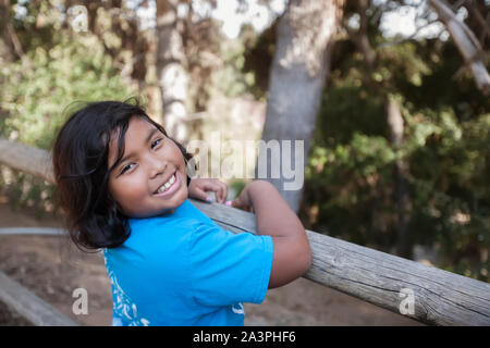 Una bambina con un grande sorriso che è tenuta su di una staccionata in legno e circondato da alberi in un ambiente naturale. Foto Stock