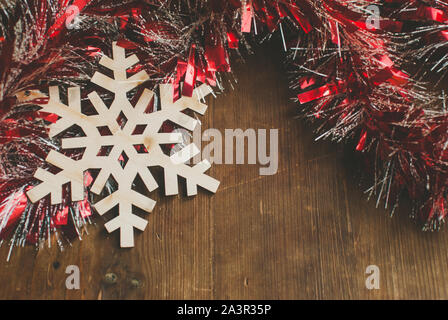 Sfondo di natale con composizione decorativa di tinsel rosso e il simbolo del fiocco di neve in legno su tavola di legno. Foto Stock