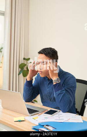 Sensazione di esaurimento. Frustrati giovane uomo barba massaggiare la sua testa e tenere gli occhi chiusi mentre è seduto al suo posto di lavoro in ufficio Foto Stock