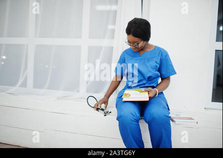 Ritratto di donna felice americano africano giovane medico pediatra in uniforme blu e il cappotto di uno stetoscopio con libri a mani. Sanitario, medico, med Foto Stock