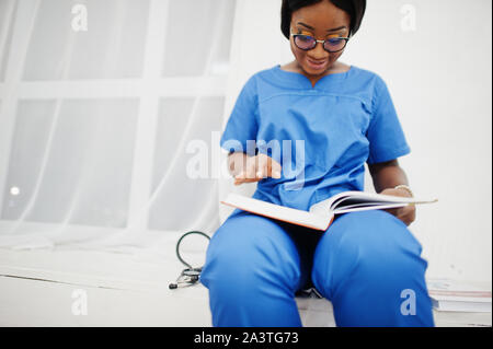 Ritratto di donna felice americano africano giovane medico pediatra in uniforme blu e il cappotto di uno stetoscopio con libri a mani. Sanitario, medico, med Foto Stock
