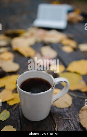 Su un vecchio tavolo di legno in autunno park è una tazza di tè e caffè, sparse in foglie di giallo e pigne. Vista dall'alto, in blur. Autunno Caldo buio Foto Stock