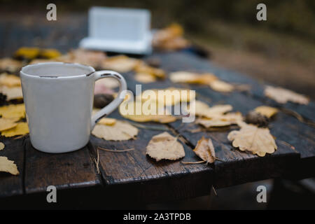 Su un vecchio tavolo di legno in autunno park è una tazza di tè e caffè, sparse in foglie di giallo e pigne. Vista dall'alto, in blur. Autunno Caldo buio Foto Stock