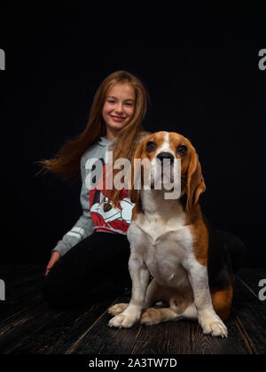 Una i Capelli rossi ragazza di dieci anni suona con un cucciolo di beagle seduto su un pavimento di legno scuro di fronte uno sfondo nero. Foto Stock