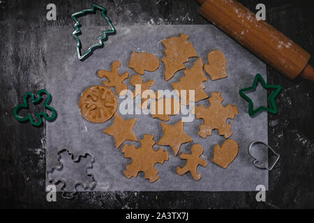 Tagliare gingerbread cookie nella forma di un albero di Natale, stella piccolo uomo, cuori da impasto crudo su pergamena carta da forno su uno sfondo scuro Foto Stock