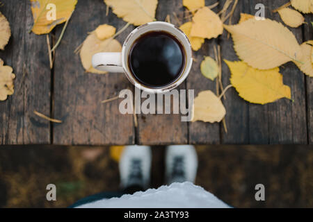 Su un vecchio tavolo di legno in autunno park è una tazza di tè e caffè, sparse in foglie di giallo e pigne. Vista superiore, gambe visibile in blur. Autunno Foto Stock