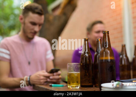 Felice l'uomo seduto e utilizzando il telefono all'aperto pur avendo la birra la messa a fuoco su oggetti in primo piano Foto Stock