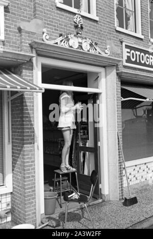 Junge Frau im Minikleid beim Fensterputz in einem Fotogeschäft a Volendam, Niederlande 1971. Giovane donna che indossa un mini dress pulizia dei vetri di un negozio fotografico a Volendam, Paesi Bassi 1971. Foto Stock