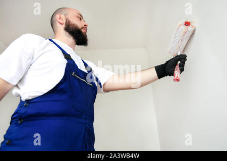 Operaio edile finitura facendo lavori di rinnovo nell'appartamento. Pittore professionista contraente utilizzando vernice bianca spazzola a rullo pittura del muro di cemento. Foto Stock