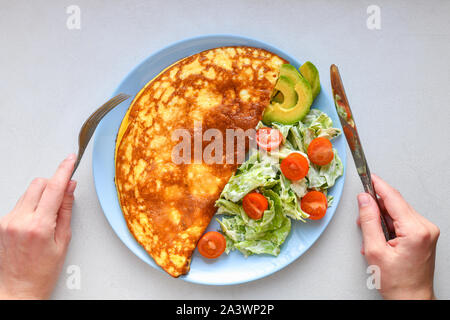 Uova fritte con verdure. le mani nel telaio. Frittata di vegetali su una piastra blu su un tavolo bianco, vista dall'alto Foto Stock