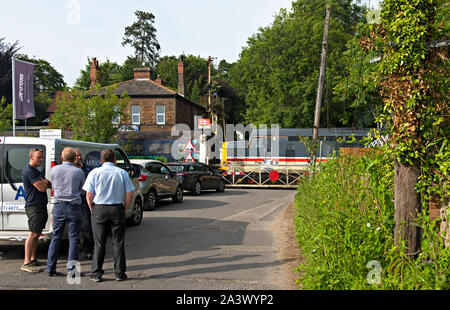 Il traffico attende al passaggio a livello Brundall stazione ferroviaria, Norfolk, Regno Unito. Una classe 37 locomotiva diesel passa la traversata. Foto Stock