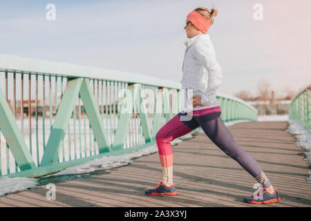 Ritratto di un sano donna bionda mantenendo montare uno una fredda giornata invernale facendo alcuni tratti prima di andare a correre su un ponte in un snowy urbano per la città Foto Stock