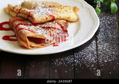 Close-up di crepes piegato in quattro sulla piastra bianca con marmellata di fragole su un tavolo di legno Foto Stock