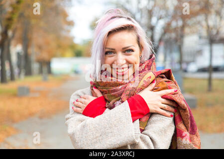 Una giovane ragazza sorride e avvolge se stessa in una sciarpa. Autunno freddo. Ritratto di una bella ragazza. Foto di strada. Foto Stock