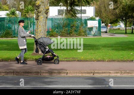 Una madre spingere un passeggino, una carrozzina o un passeggino per la strada mentre guarda al suo telefono cellulare o telefono cellulare Foto Stock