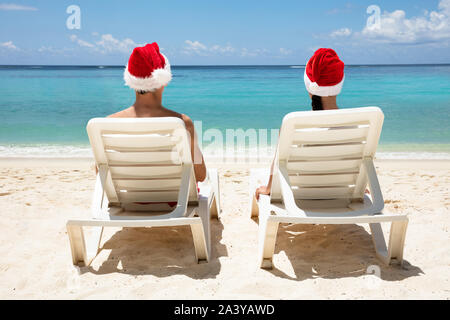 Vista posteriore di un paio di indossare cappelli di Babbo Natale rilassante su bianco Sedie a sdraio sulla spiaggia Foto Stock