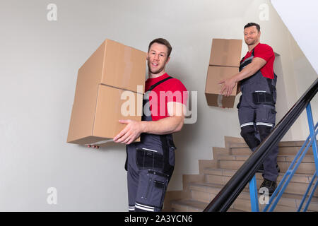 Ritratto di un Giovane Maschio Movers in uniforme che porta le scatole di cartone a piedi verso il basso sulla scala Foto Stock