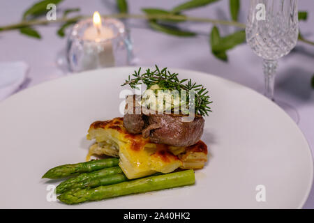 Piastra di colore bianco con il filetto di angus, Pommes Boulangerre, asparagi, rosmarino, germogli di soia, giovani foglie di bietola. Candela accesa sullo sfondo. Vista laterale Foto Stock