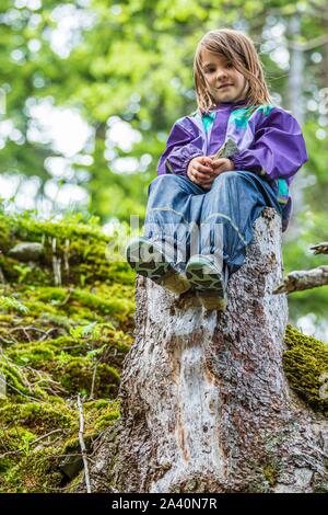 Poco ragazza seduta su di un ceppo di albero nella foresta, Safiental, Grigioni, Svizzera Foto Stock