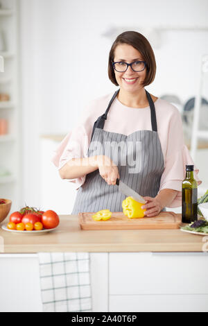 Ritratto di giovane donna in occhiali e grembiule sorridente in telecamera in posizione eretta e tagliare le verdure in cucina a casa Foto Stock