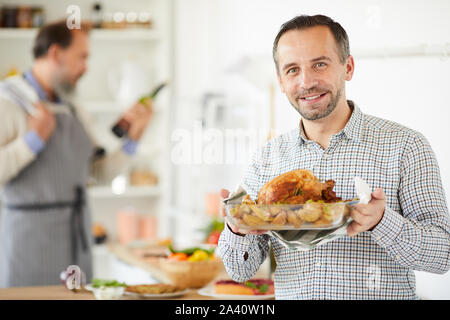 Ritratto di giovane uomo con piatto di pollo arrosto e sorridente alla telecamera mentre un altro uomo preparare il cibo in background Foto Stock