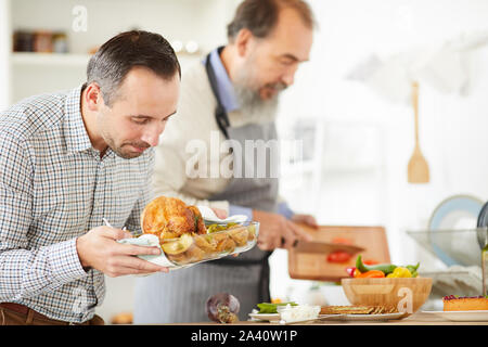 Giovane uomo che guarda il tacchino arrosto nelle sue mani con i senior uomo vegetale di taglio in background a casa Foto Stock