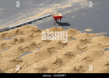 Carriola rossa con una ruota sul marciapiede e molti piccoli scivoli di sabbia sulla terra. Migliorare i percorsi su strada nella città, il lavoro al di fuori su un caldo sunn Foto Stock