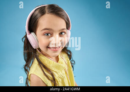 Bambino positivo indossare gli auricolari rosa guardando la fotocamera e ridere sul blu sfondo isolato. Graziosa bambina in maglietta gialla ascoltando musica e godendo di dispositivo moderno. Concetto di tecnologia. Foto Stock