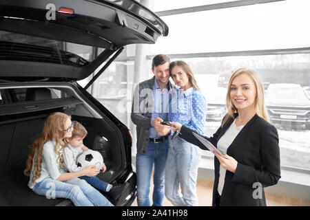 Vista dal lato della femmina venditore auto guardando la fotocamera e sorridere mentre che dà le chiavi della macchina a nuovi proprietari in auto salon. Concessionaria auto vendita di nuove auto per la famiglia felice. Concetto di acquisto automatico. Foto Stock