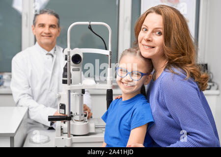 Vista dal lato di felice la madre e il bambino controllo visivo in clinica. Famiglia allegra guardando la fotocamera e sorridente insieme mentre medico seduto a sfondo. Concetto di Cura degli occhi e della vista. Foto Stock