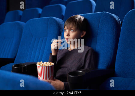 Vista dal lato del ragazzo eccitato con grandi occhi e bocca aperta a guardare fantastico film per il cinema. Scioccato teen maschio mangiare popcorn, guardando lo schermo e la visione di orrore. Concetto di svago e divertimento. Foto Stock