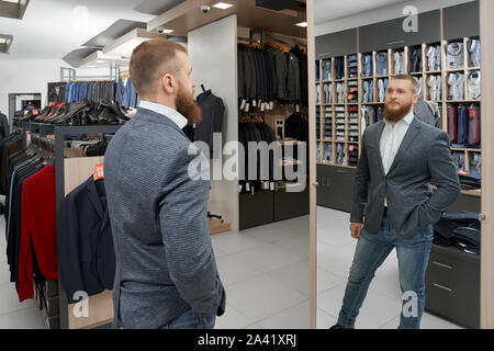 Giovane gentiluomo in elegante giacca casual in piedi di fronte ad uno specchio e guardando la riflessione in boutique. Client maschio cercando su e acquisto di soddisfare nel grande negozio di abbigliamento. Concetto di vestito e di montaggio. Foto Stock