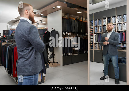 Grave client maschio guardando lo specchio, scelta e acquisto di nuovi smart casual muta in negozio. Giovane imprenditore cerca sulla camicia alla moda nella boutique. Concetto di stile e di abbigliamento maschile. Foto Stock