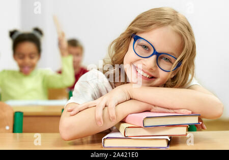 Vista frontale della bella femmina studente guardando la fotocamera e ridere mentre giacenti sui libri in aula. Smart girl in vetri appoggiato su apparecchiature, in posa e sorridente a scuola. Concetto di apprendimento. Foto Stock