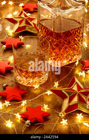 Crystal decanter di whisky e un bicchiere di whisky con le luci di Natale e stelle rosse decorazioni, su di un tavolo di legno Foto Stock