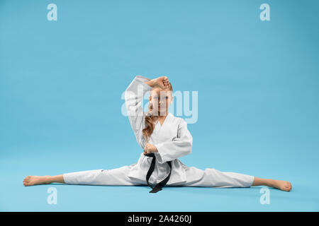 Fiducioso ragazza in kimono bianco con cintura nera seduto sulla croce lo spago e guardando la fotocamera. La formazione del bambino stretching e posa su sfondo blu. Foto Stock