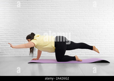 Giovane donna con la curva del corpo e capelli castani knot stare su un ginocchio sul tappetino viola e stirando la sua mano sinistra e gamba destra. Paffuto femmina in felpa con cappuccio giallo e nero leggings esercita in ambienti interni Foto Stock