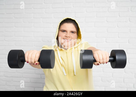 Sorridente brunette chunky giovane donna in felpa con cappuccio giallo guardando la fotocamera e trattenere due big black dumbbell sulle sue mani con muro bianco dietro.donna grassa è seriamente impostato per portare la stessa in buona forma Foto Stock