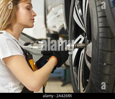 Vista laterale della bellissima femmina cambio meccanico pneumatico di automobile in officina. Bionda in maglia bianca, tuta e guanti di protezione lavora in auto service. Concetto di manutenzione e riparazione. Foto Stock