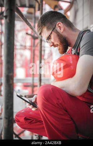 Sito di costruzione lavoratore seduto ad alta sul ponteggio, riposo e utilizzando il telefono cellulare Foto Stock