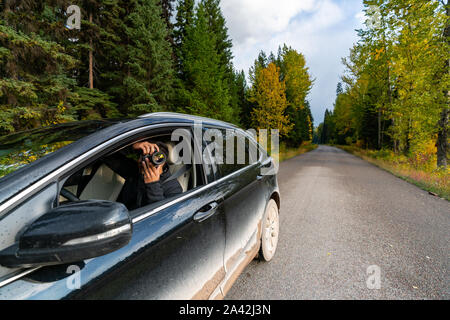 Uomo di fotografare la natura dall'interno del suo sporco nero auto e gli alberi con caduta calors in background, Montana. Foto Stock